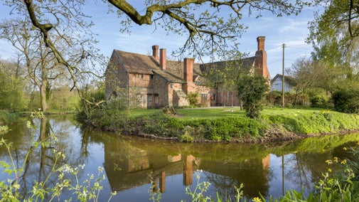 A view across a moat to the back of the manor house at Brockhampton on a sunny day, with the house reflected in the water